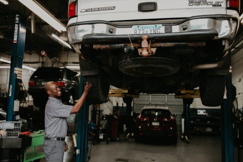 Mechanic smiling while inspecting under a raised SUV in an auto repair shop.