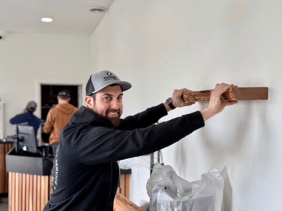 Man installing a wooden shelf on a white wall in an office setting, smiling at the camera.