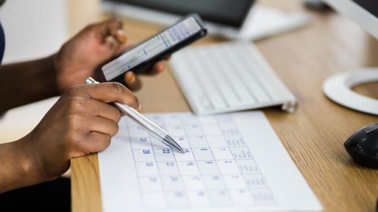 Person planning schedule with calendar and smartphone, sitting at desk with keyboard and mouse.