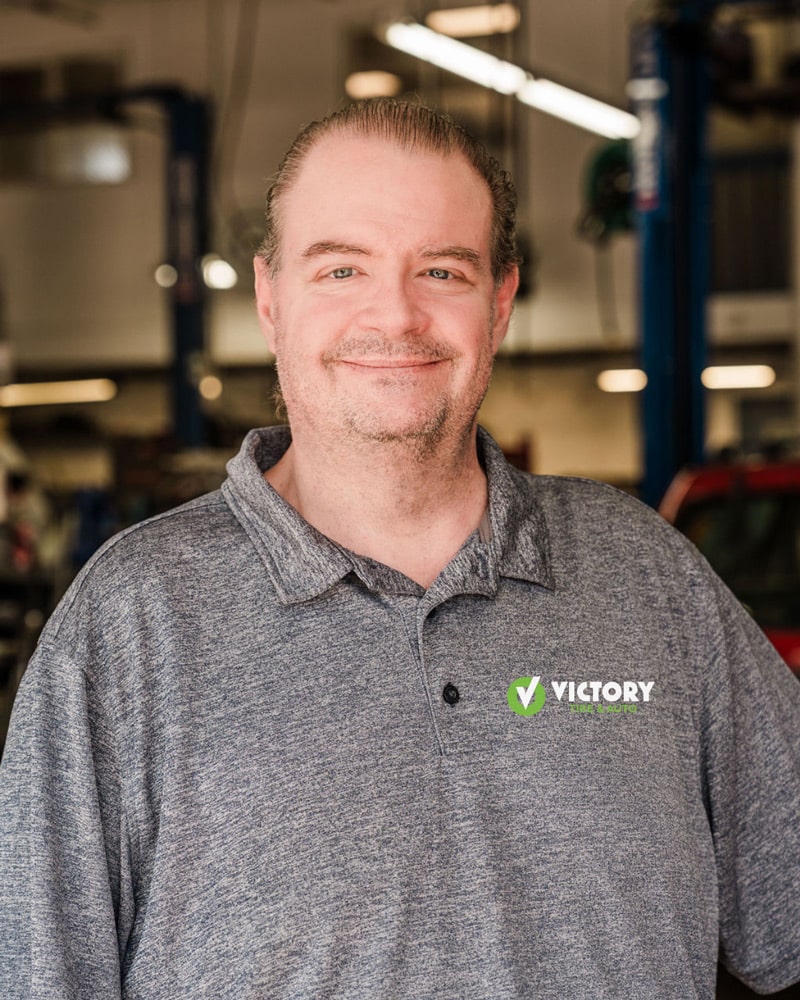 Smiling person in a gray shirt at Victory Tire & Auto, with a garage background.