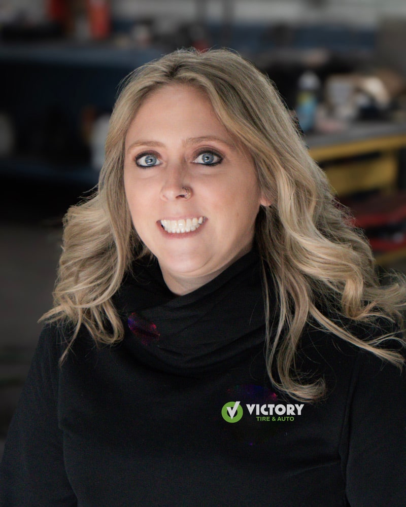 Smiling woman in a black Victory Tire & Auto shirt, standing in an auto shop environment.