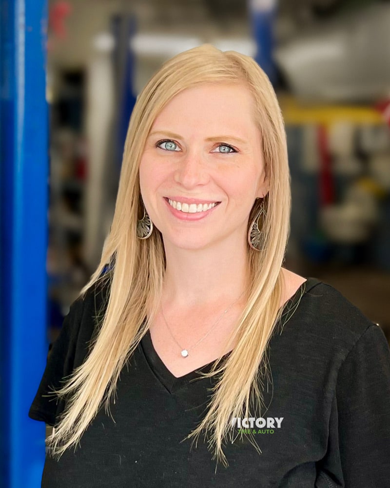 Smiling woman at Victory Tire & Auto shop, wearing a black shirt with company logo, indoors.