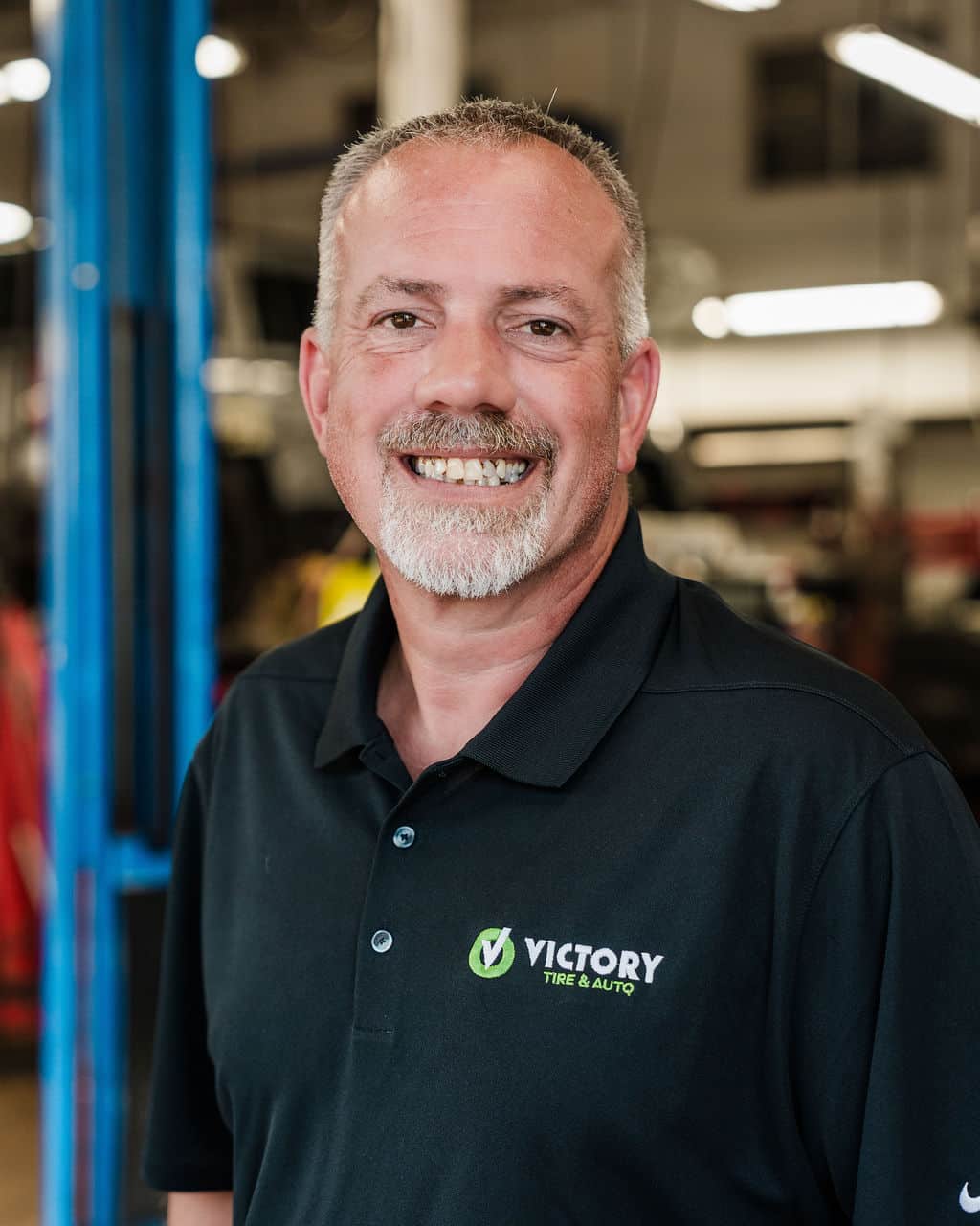 Smiling man in a Victory Tire & Auto shirt, standing in a garage with a friendly expression.