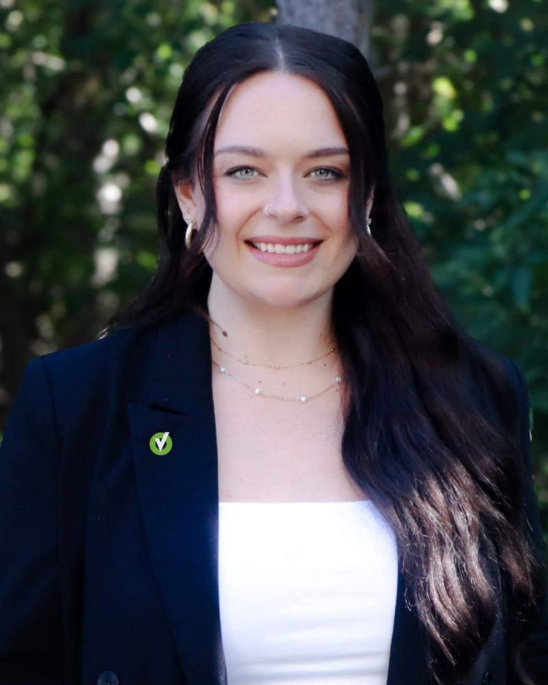 Smiling woman with long dark hair in a blazer, outdoors with greenery in the background.