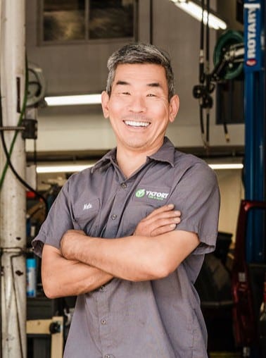 Smiling mechanic in a garage with arms crossed, wearing a gray uniform shirt, standing by automotive equipment.