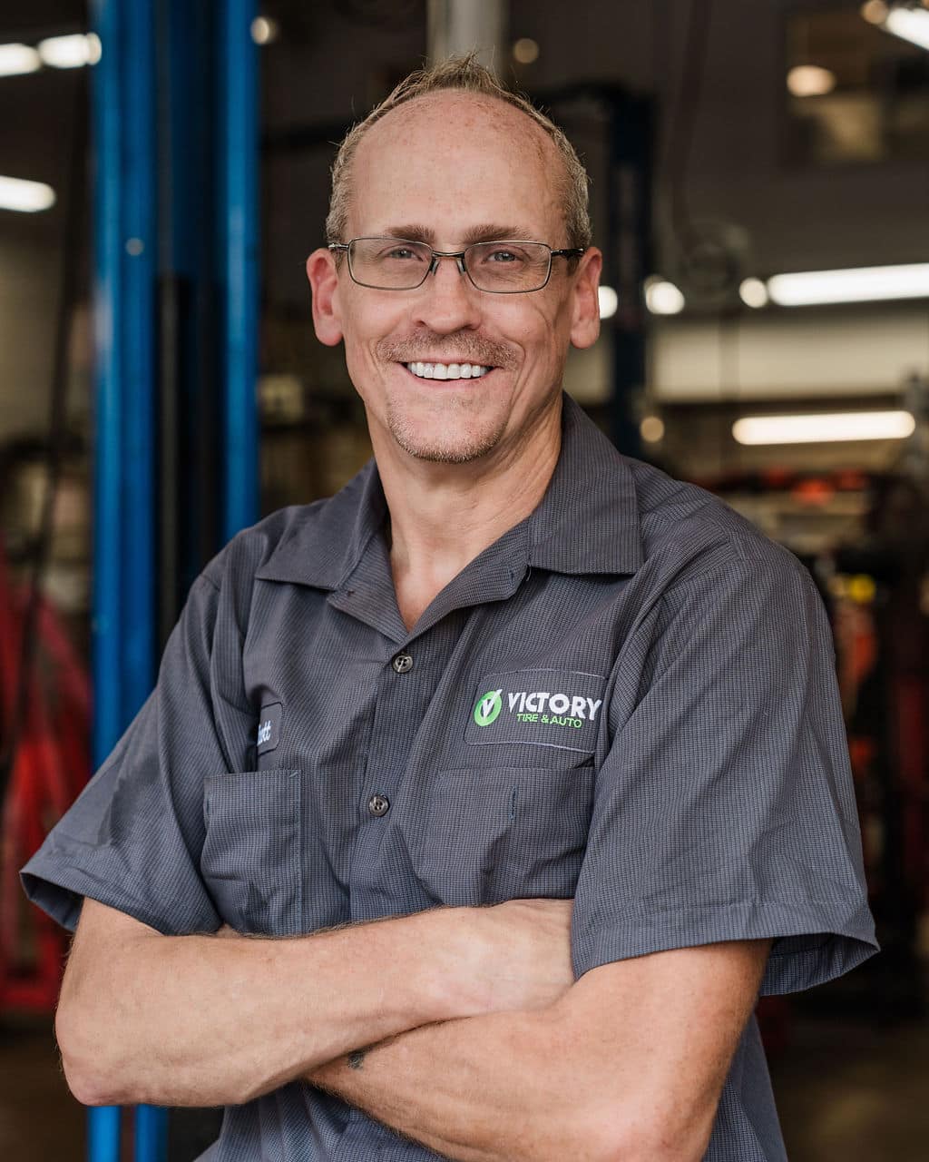 Smiling mechanic in a workshop wearing Victory Tire & Auto uniform with arms crossed.